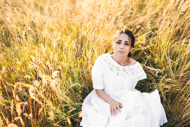 Beautiful girl in white dress sitting in wheat field at sunset - Zdjęcie, obraz