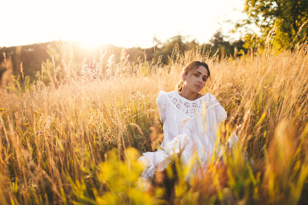 Beautiful girl in white dress sitting in wheat field at sunset - Фото, изображение