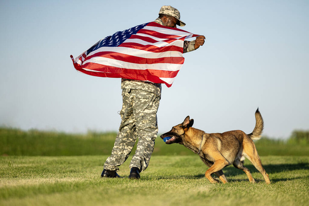 Katonai egyenruhás katona, amerikai zászlóval a kezében és a kutyájával játszik. Büszkék az országukra.. - Fotó, kép