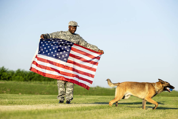 Katona egyenruhában, amerikai zászlót tartva, a függetlenség napját ünnepli a katonai kutyájával.. - Fotó, kép