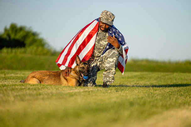 Dumni ze swojego kraju. Żołnierz pokryty flagą USA bawiący się psem wojskowym na trawie. - Zdjęcie, obraz