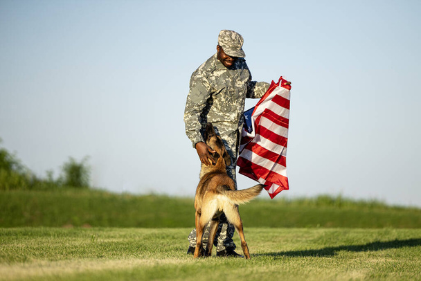Soldat und Militärhund spielen auf dem Feld und halten die US-Flagge hoch. Nationalstolz. - Foto, Bild