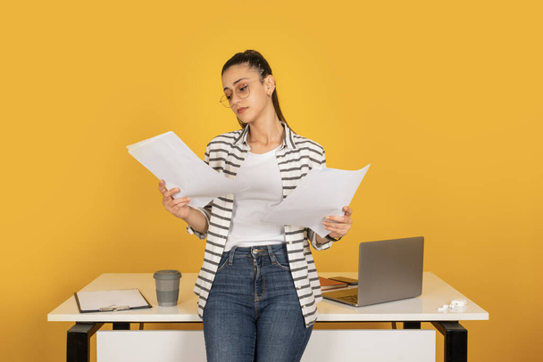 Checking account documents, portrait of young successful business woman checking account documents. Leaning work office desk holding papers. Isolated yellow background. Career lifestyle concept idea. - Photo, Image