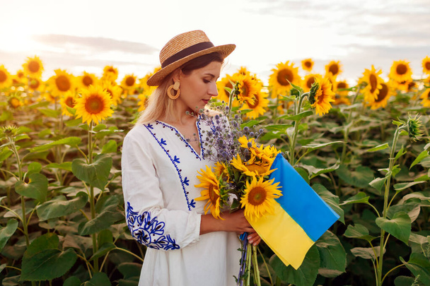 Молодая женщина с желто-голубым флагом Украины с подсолнухами на летнем поле в национальной одежде с вышивкой. День независимости Украины. Символ свободы и мира - Фото, изображение