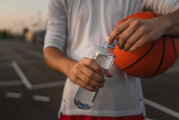 close up on hands midsection of unknown caucasian man teenager open plastic bottle of water outdoor in sunny day doing sports with basketball - Photo, Image