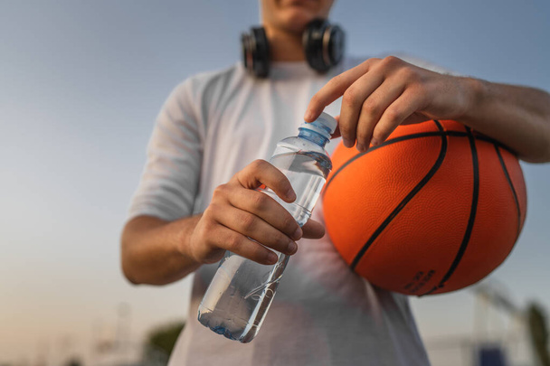 primer plano en las manos sección media de hombre caucásico desconocido adolescente abierto botella de plástico de agua al aire libre en día soleado haciendo deportes con baloncesto - Foto, Imagen
