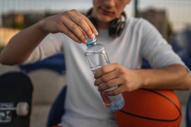 primer plano en las manos sección media de hombre caucásico desconocido adolescente abierto botella de plástico de agua al aire libre en día soleado haciendo deportes con baloncesto - Foto, imagen