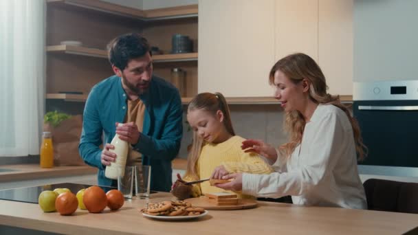 Valkoihoinen onnellinen perhe keittiössä aamulla aamiainen ruoan jakelupalvelulla vanhemmat äiti isä lapsen kanssa tytär lapsi tyttö ruoanlaitto paahtoleipää suklaalla pasta isä kaada maitoa lasiin. - Materiaali, video