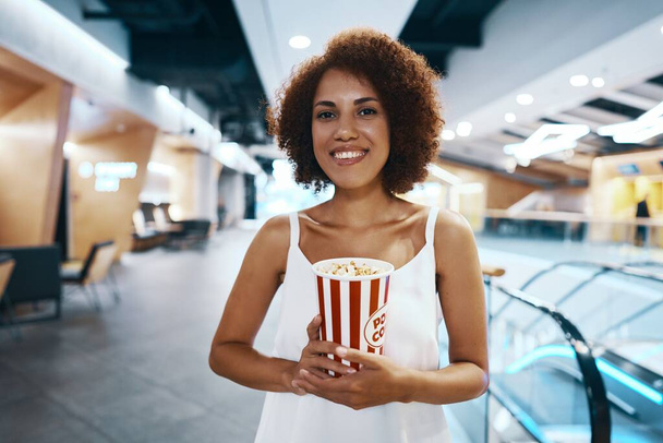 Curly-haired woman with an afro hairstyle in a white shirt is walking to watch a movie with popcorn in hand. High quality photo - Fotografie, Obrázek