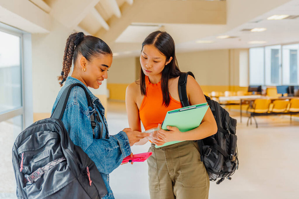Két fiatal nő, egy latin és egy ázsiai osztálytárs okostelefont használ a közösségi hálózatok ellenőrzésére vagy multimédiás tartalmak megtekintésére a középiskolájuk vagy egyetemük folyosóin - Fotó, kép
