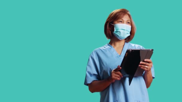 Happy clinic nurse looking at patient data on digital tablet, confused with clinical checkup results, close up. Experienced healthcare specialist wearing face mask, isolated over studio background - Footage, Video