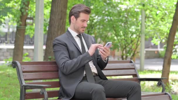 Mladý podnikatel slaví úspěch na chytrém telefonu, zatímco sedí venku na lavičce - Záběry, video