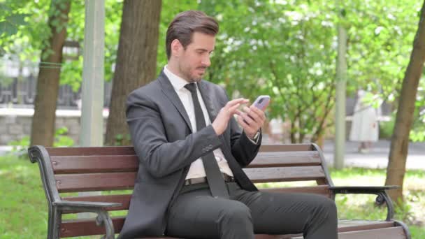Fiatal üzletember sokkolta a veszteség a Smartphone miközben ül a szabadban egy padon - Felvétel, videó