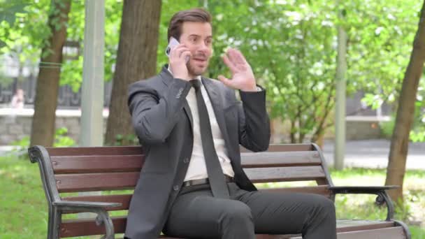 Молодий підприємець сперечається по телефону сидячи на відкритому повітрі на лавці - Кадри, відео