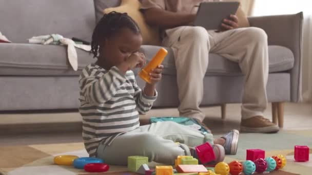 Cute 3 year old African American girl playing with toys on rug in living room while staying at home with dad sitting on couch with digital tablet in hands - Footage, Video