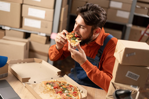 Männliche Lagerarbeiter essen mittags oder abends leckere Pizza, während sie am Arbeitsplatz sitzen. Hoher Winkel - Foto, Bild