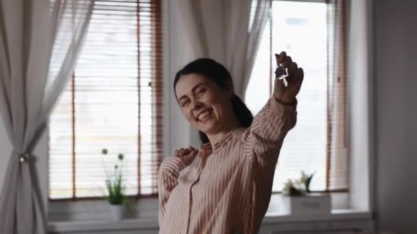 Vermietung und Kauf von Immobilien. Porträt einer glücklichen kaukasischen Frau, die tanzt und Schlüssel aus ihrer neuen Wohnung vorzeigt. Fenster im Hintergrund. Hypotheken- und Kreditkonzept. - Filmmaterial, Video