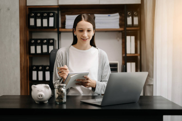 happy smiling financial business woman making notes at office desk with piggy bank, money coins laptop and calculator - Foto, imagen