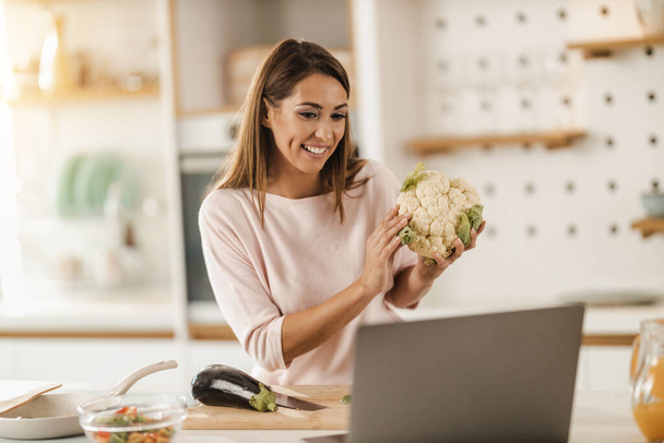 Egy fiatal, mosolygós nőről készült felvétel, aki egészséges ételt készít, és videóblogot készít egy laptopon otthon.. - Fotó, kép