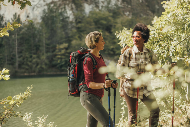 Due amiche di razza mista in piedi accanto a un lago che fanno una pausa durante un'escursione nella natura. - Foto, immagini