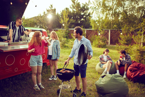 Group of young people, friends spending time outdoors, having barbecue party, cooking, drinking cocktails, talking. Countryside meeting. Concept of friendship, leisure time, weekends, summer, party - Photo, Image