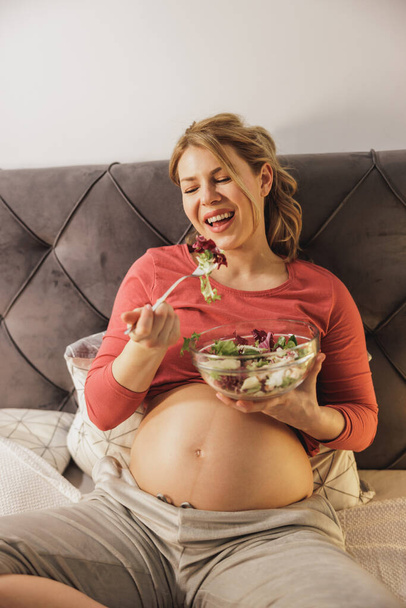 Feliz mujer embarazada comiendo ensalada de verduras frescas mientras está sentada en la cama en su dormitorio. - Foto, Imagen