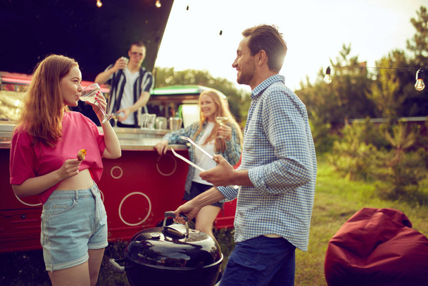 Young smiling man and woman cooking meat, doing barbecue, drinking cocktails and talking. Blurred people on background. Good time outdoors. Concept of friendship, leisure time, weekends, summer, party - 写真・画像