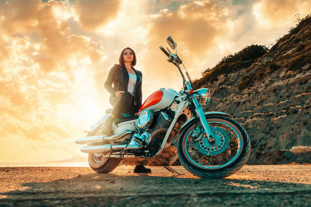 Piękna pewna siebie dorosła kobieta w skórzanych ubraniach siedząca na zabytkowym motocyklu. Widok na dół. Epicki zachód słońca na tle. Koncepcja podróży motocyklowych i feminizmu. - Zdjęcie, obraz