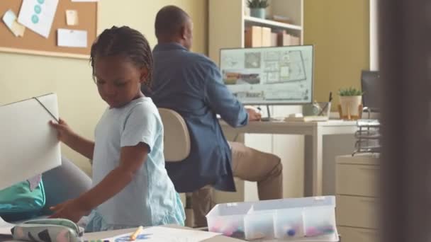 Musta mies arkkitehti kehittää talon suunnitelma tietokoneella toimistossa, kun hänen kaunis 3-vuotias tytär piirustus sohvapöytä lähellä - Materiaali, video