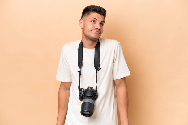 Joven fotógrafo caucásico aislado sobre fondo beige haciendo gestos de dudas mientras levanta los hombros - Foto, Imagen