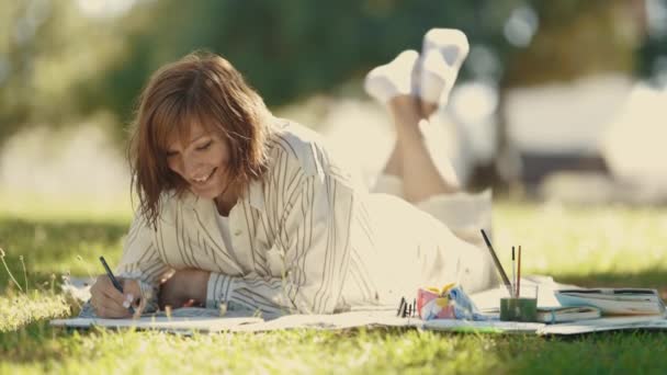 Una mujer adulta acostada en la manta en el parque floreciente y dibujando en el cuaderno de bocetos. Mid shot - Metraje, vídeo