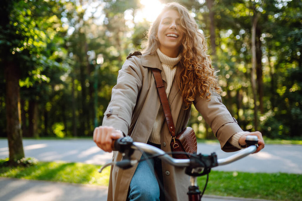 Happy pensive young woman in stylish clothes riding a bike in a sunny park, outdoors, looking away. Active lifestyle. The concept of nature. - Foto, Imagen