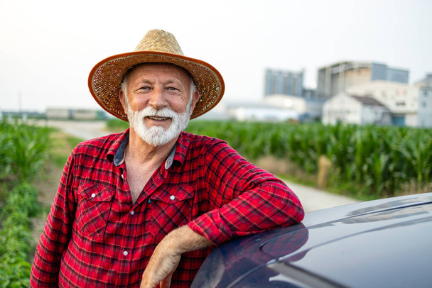 Portret starszego rolnika stojącego przy polu i opierającego się na maszynie ciągnika. - Zdjęcie, obraz