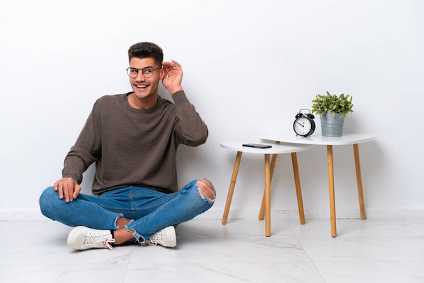Fiatal kaukázusi férfi ül otthonában elszigetelt fehér háttér hallgat valamit azáltal, hogy kezét a fülre - Fotó, kép