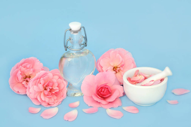 Rozenbloem parfum in hartvormige fles met bloemen en losse bloemblaadjes op blauw. Voorbereiding van geparfumeerd bloemproduct, cadeau voor Valentijnsdag, verjaardag, jubileum of Moederdag. - Foto, afbeelding