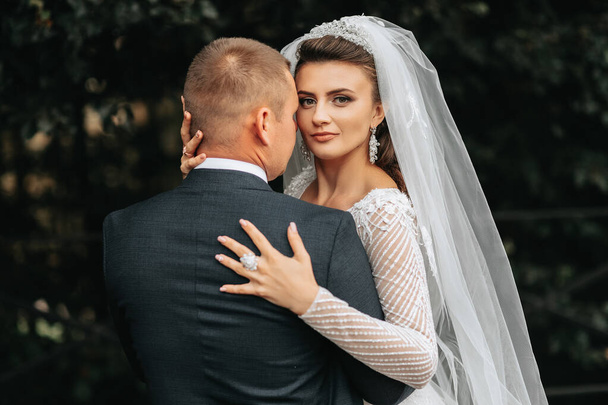 Wedding portrait. A brunette bride in a long dress and a groom in a classic suit are standing, hugging, in love and posing for the camera. Beautiful bride, lace dress - Zdjęcie, obraz