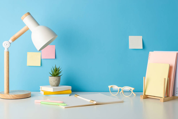 Academic essentials, such as books holder, copybook, glasses, and lamp neatly arranged on desk, captured in side view photo against blue wall with sticky notes. Perfect for text or promotional content - Foto, immagini