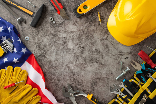 Commemorating dedication of construction workers on Labor Day. High-angle shot of flag, helmet, gloves and toolkit on grunge-textured grey concrete surface, with copyspace for text or advertisements - 写真・画像