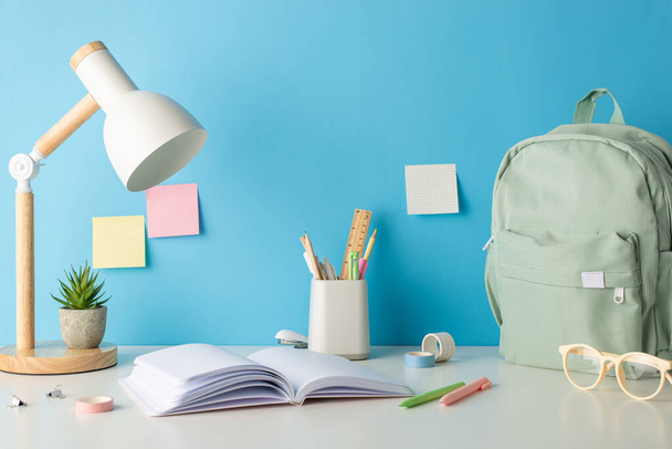 Get set for learning with this side view photo of desk equipped with school essentials, book, pencil stand, plant, glasses, backpack and more. Blue wall backdrop with sticky notes ideal for text or ad - Photo, Image