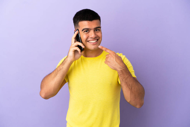 Fiatal jóképű férfi használja a mobiltelefon felett elszigetelt lila háttér ad egy hüvelykujj fel gesztus - Fotó, kép