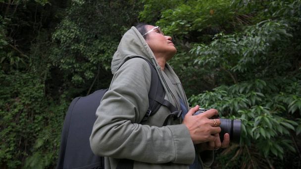 Solo Asyalı kadın doğa ciddi fotoğrafçı kamera aygıtı tutuyor yemyeşil yaprak yağmur ormanlarında çarpıcı görüntüler arıyor. Açık hava aktiviteleri. Doğada güzellik. Seyahat hedefi. - Fotoğraf, Görsel