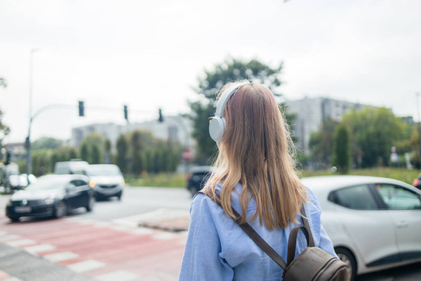 Vista trasera de la mujer joven con auriculares mujer escuchando la música, cruzando la calle en los semáforos. Foto de alta calidad - Foto, Imagen