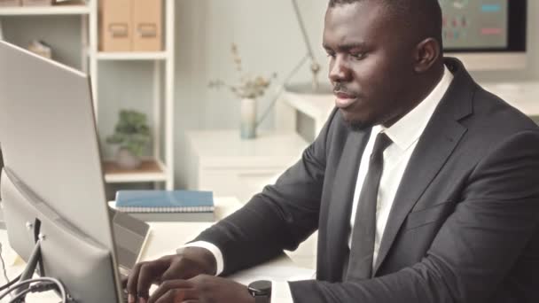 Enfocado joven hombre de negocios afroamericano en elegante traje negro escribiendo en el teclado inalámbrico mientras se trabaja en PC en la oficina moderna - Imágenes, Vídeo