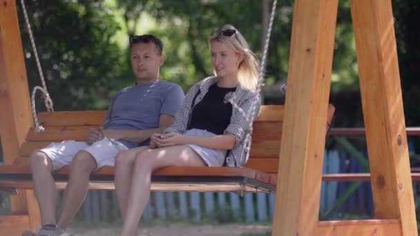 Young couple man and woman riding on wooden swing in outdoor park. Guy strokes girl's hair . Married couple in love enjoys. Young family and lovers. - Footage, Video