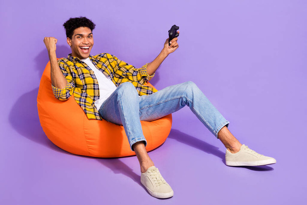 Full body photo of delighted positive person sit comfy bag hold controller raise fist isolated on violet color background. - 写真・画像