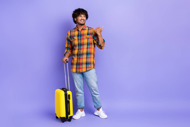 Foto corpo pieno di giovane ragazzo indossare jeans alla moda diretta dito mockup programma aeroporto portare valigia isolata su sfondo di colore viola. - Foto, immagini