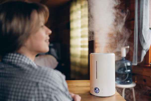 A modern air humidifier during relaxation or rest, a happy young woman, a girl who enjoys the aromatherapy steam aroma from a diffuser with essential oils, feels comfortable in a room, apartment, house. - Photo, Image