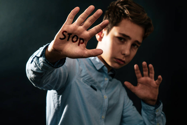 The concept of domestic violence. Sad teenager puts his hands in front of him defending himself from the threat of attack. Black background. Text STOP . - Photo, image