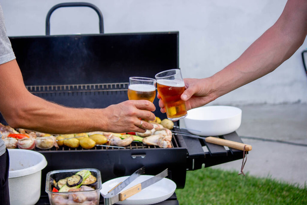 mannen houden bier in hun handen op de achtergrond van de grill, groenten en vlees. Selectieve focus. Natuur - Foto, afbeelding