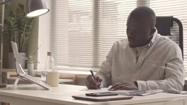 オフィスの机で財務報告をしながらノートパソコンを使用している深刻なアフリカ系アメリカ人男性 - 映像、動画